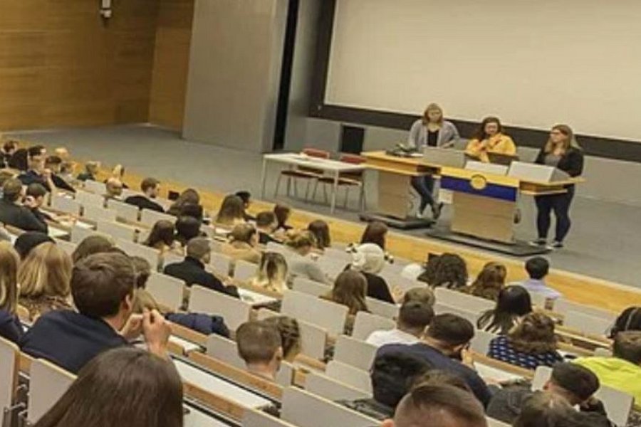 Ein gefüllter Vorlesungssaal. Man schaut aus der Sicht der Zuhörenden. Vorne stehen 3 Personen an einem Pult.