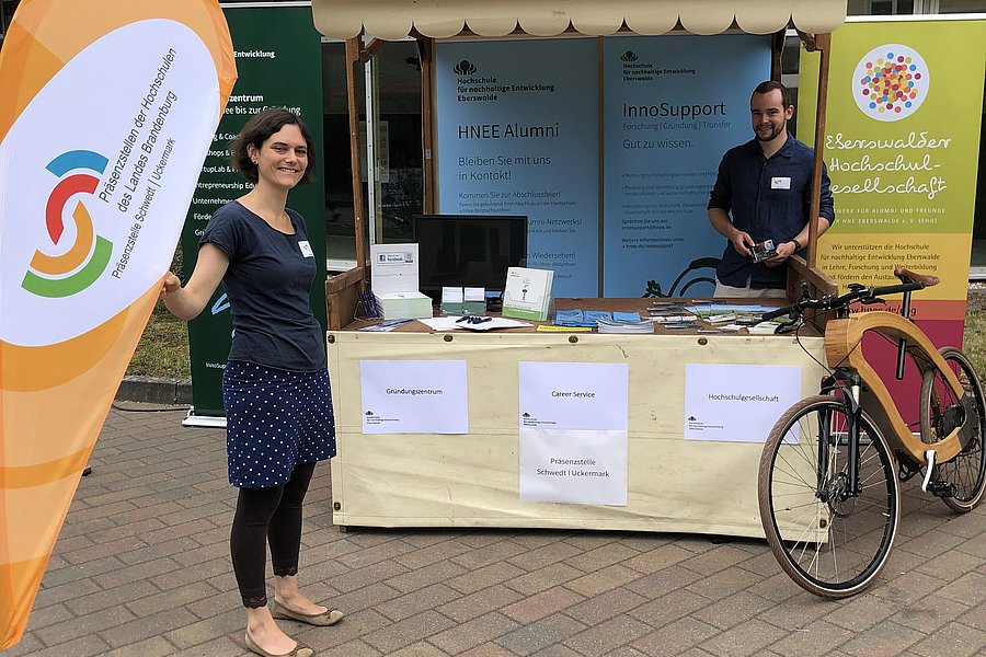 In der Mitte befindet sich der Messestand der Präsenzstelle Schwedt l Uckermark zum Tag der offenen Tür. Ein Mitarbeiter steht dahinter. Links im Bild ist die Beachflag der Präsenzstelle zu sehen und daneben eine weitere Mitarbeiterin der Präsenzstelle. 