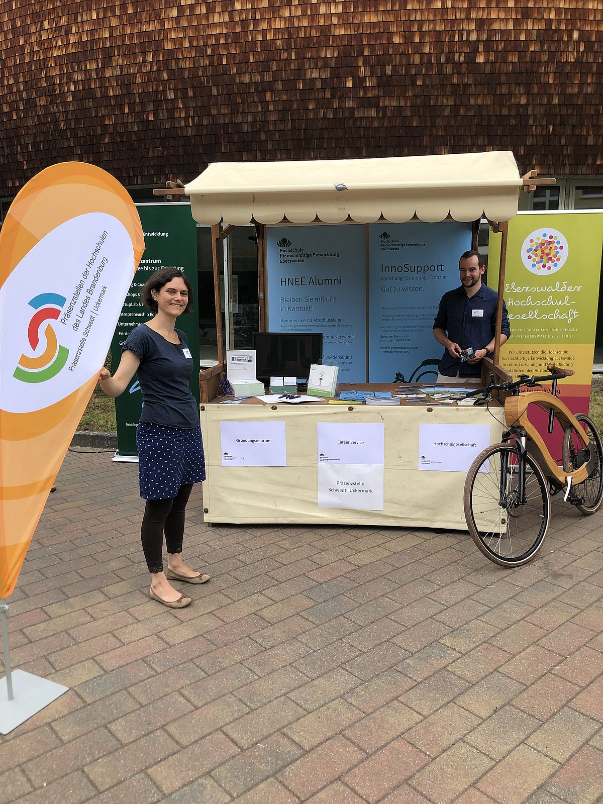 In der Mitte befindet sich der Messestand der Präsenzstelle Schwedt l Uckermark zum Tag der offenen Tür. Ein Mitarbeiter steht dahinter. Links im Bild ist die Beachflag der Präsenzstelle zu sehen und daneben eine weitere Mitarbeiterin der Präsenzstelle. 