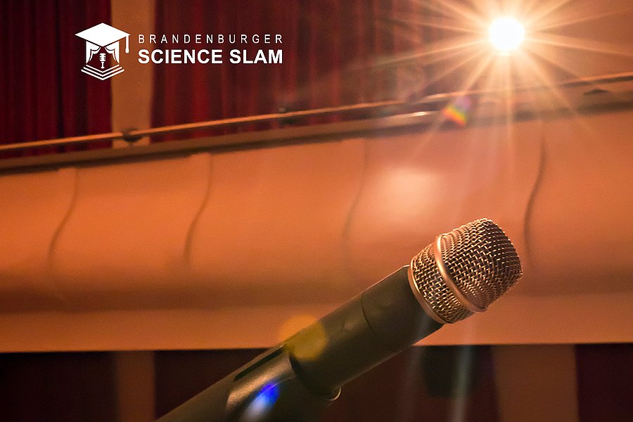 Im Vordergrund ein Mikrofon. Ein Scheinwerfer ist darauf gerichtet. Im Hintergrund erkennt man den leeren Zuhörersaal. Oben links in der Ecke befindet sich der Schriftzug "Brandenburger ScienceSlam" und das Logo dazu.