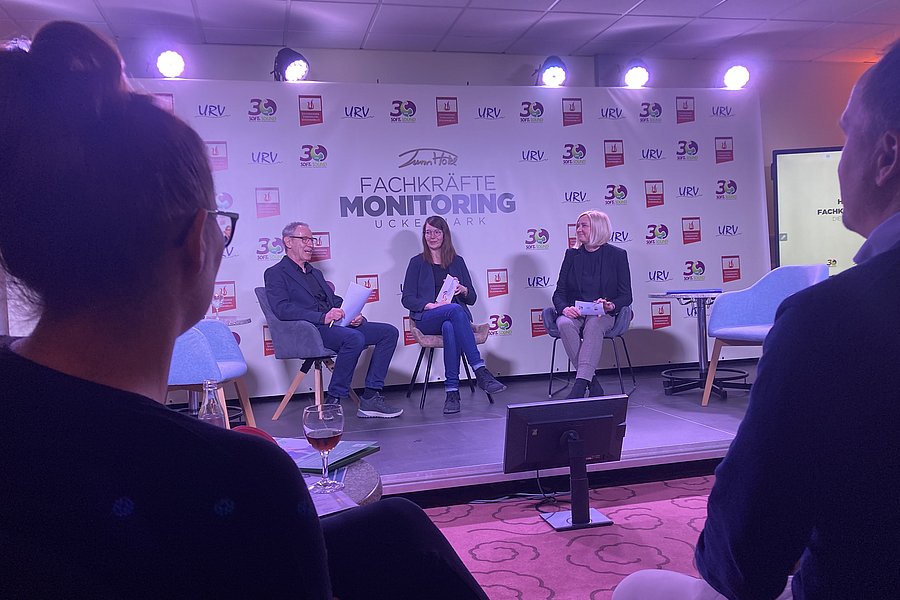 Aus der Zuschauerperspektive schaut man auf ein Podium. Dort sitzen drei Personen für eine Diskussion. Im Hintergrund erkennt man den Schriftzug "Fachkräftemonitoring Uckermark". Im Vordergrund sind links und rechts zwei Zuhörende von hinten zu erkennen."