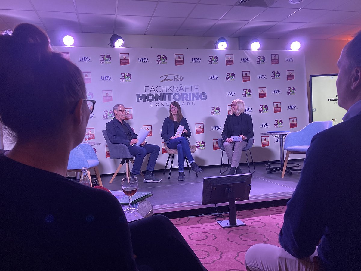 Aus der Zuschauerperspektive schaut man auf ein Podium. Dort sitzen drei Personen für eine Diskussion. Im Hintergrund erkennt man den Schriftzug "Fachkräftemonitoring Uckermark". Im Vordergrund sind links und rechts zwei Zuhörende von hinten zu erkennen."