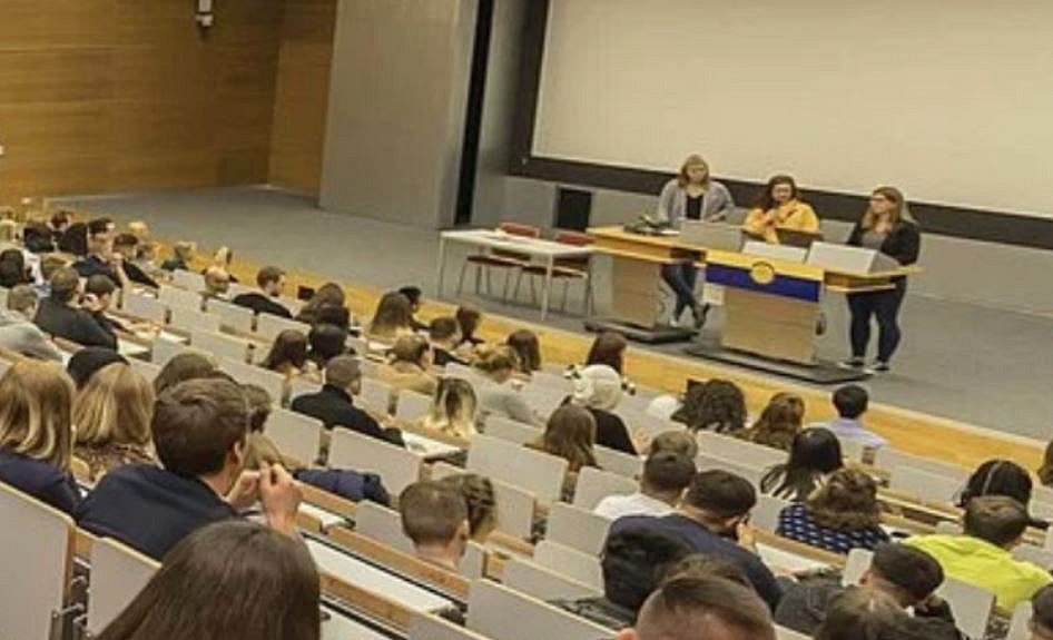 Ein gefüllter Vorlesungssaal. Man schaut aus der Sicht der Zuhörenden. Vorne stehen 3 Personen an einem Pult.